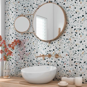 May include: A modern bathroom scene featuring a white oval sink, gold fixtures, and a round mirror. The wall is decorated with a terrazzo pattern in various colors. A vase of red flowers adds a pop of color to the wooden countertop.