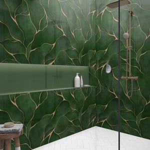 Op de afbeelding: Een moderne badkamer met groene muren met een bladmotief in goud. De doucheruimte heeft een glazen deur, een bronzen douchekop en witte tegels in baksteenstijl. Een kleine houten kruk en een plank met witte flessen zijn ook zichtbaar.