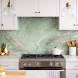 May include: A bright kitchen with white cabinets, gold hardware, and a green marble backsplash. A stainless steel gas range is centered below the backsplash. A loaf of bread, wooden utensils, and a basket of lemons are visible.