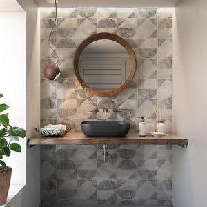May include: A modern bathroom with a black oval sink, a wooden countertop, and a round wooden mirror. The wall features a geometric tile pattern in shades of gray. A small pendant light hangs above the sink, and a potted plant sits in the corner.