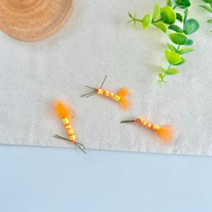 May include: Three handmade fishing flies with yellow and orange accents. Each fly features a segmented yellow body, orange feather tail, and thin wire hooks. The flies are arranged on a white surface with green foliage and a wooden disc in the background.