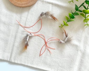 May include: Three cat toys with gray and brown feathers and red string. The toys are arranged on a white cloth with a wooden circle and green plant in the background. The toys are designed to entice cats to play.