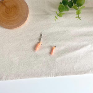 Könnte beinhalten: Ein Paar orangefarbene und schwarze Ohrringe, einer mit einer silbernen Kette und einem Verschluss. Die Ohrringe sind wie kleine, stilisierte Karotten geformt. Die Ohrringe sind vor einem weißen Hintergrund mit einer Holzscheibe und einer grünen Pflanze platziert.