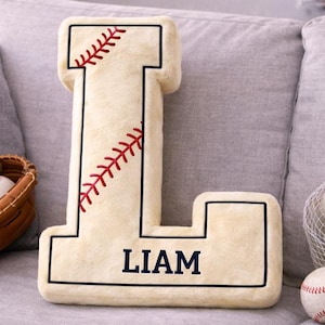 May include: A beige baseball-themed pillow shaped like the letter "L" with the name "LIAM" in navy blue. The pillow features red stitching details and is surrounded by baseballs, a glove, and a bat.