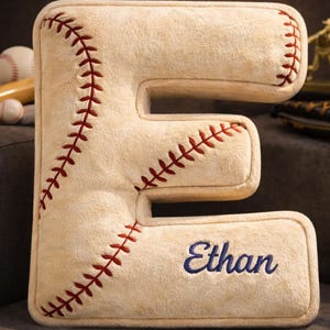 May include: A beige plush letter "E" cushion with red stitching, designed to look like a baseball. The name "Ethan" is embroidered in blue. Baseball-themed items are in the background.