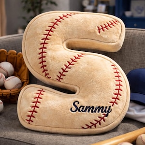 Könnte beinhalten: Beiges Baseball-Kissen in Form des Buchstabens "S" mit roten Nähten und dem Namen "Sammy" in Marineblau gestickt. Baseballhandschuh, Bälle und ein Baseballschläger sind ebenfalls zu sehen.