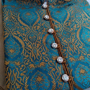 May include: Teal and gold brocade fabric with an ornate floral pattern. The garment features a mandarin collar and is fastened with decorative silver buttons. The trim is a contrasting brown color. The fabric is folded, showing the texture and design.
