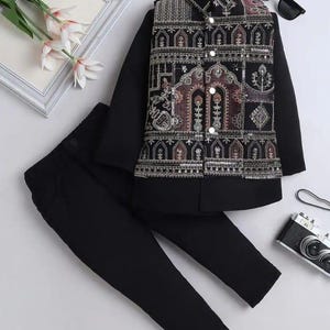 May include: A smart black outfit for a child, comprising a long-sleeved shirt, trousers, and a decorative waistcoat. The waistcoat has an elaborate embroidered pattern in brown, gold, and silver. The outfit is accessorised with sunglasses and a toy camera.