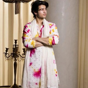 May include: A white kurta set with a vibrant tie-dye pattern in pink and yellow. The long-sleeved top features a mandarin collar and is paired with matching white pants. A white scarf is draped around the neck.