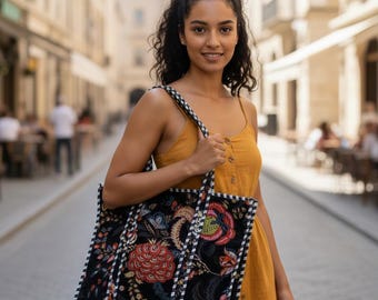 May include: A black tote bag with a colorful floral pattern, carried by a person wearing a mustard-colored dress. The bag has a white and black striped border and straps. The background is a European street scene.