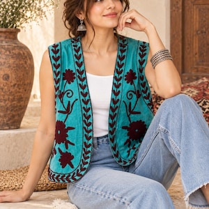 May include: A sleeveless turquoise waistcoat featuring dark maroon floral and leaf embroidery along the front edges and hem. The waistcoat is worn over a simple white vest top and paired with light blue denim jeans, creating a layered outfit.
