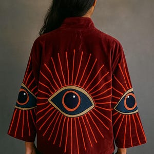 May include: A burgundy velvet jacket with embroidered eye designs. The jacket features a high collar and wide sleeves. The eye designs are in shades of orange, navy blue, and gold, with radiating lines.