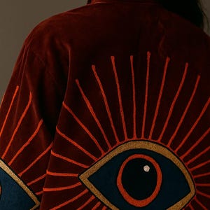 May include: A burgundy velvet jacket with embroidered eye designs. The eyes are navy blue with gold trim and a red iris, surrounded by radiating orange lines. The jacket is a rich, dark color.