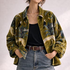 Puede incluir: Chaqueta de terciopelo verde oliva con estampado de selva con tucanes y leopardos. La chaqueta tiene cuello y cierre de botones. La modelo lleva un top negro, vaqueros azules y un cinturón marrón.