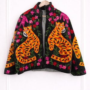 Peut inclure: Veste noire ornée de broderies de tigres orange, de fleurs roses et rouges et de feuilles vertes. La veste a un col montant et un intérieur rayé. Les tigres ont des rayures noires et sont assis. La veste est suspendue à un cintre en bois.