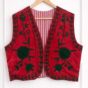 May include: A red velvet vest with intricate green floral embroidery. The vest features a striped lining and is displayed on a wooden hanger against a white background. The embroidery includes stylized flowers and leaf patterns.