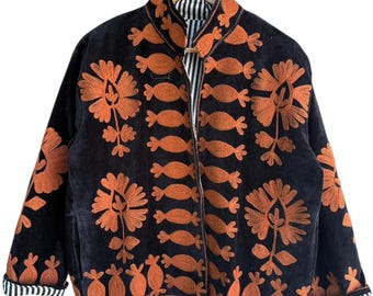 Puede incluir: Chaqueta negra con bordados florales naranjas. La chaqueta tiene cuello mandarín, mangas largas y forro a rayas. El bordado incluye flores estilizadas y diseños en forma de caramelo. La chaqueta está colgada en una percha de madera.