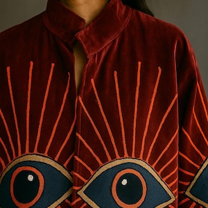 May include: A burgundy velvet jacket with orange embroidered sunburst designs radiating from large, embroidered blue and gold eye motifs. The jacket has a high collar and a relaxed fit, suitable for layering.