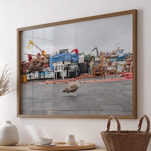 May include: A framed photograph of a harbor scene with a seagull in the foreground. The image shows fishing boats, nets, and various items on a gray paved area. The frame is a warm brown color, and the photograph is displayed above a wooden cabinet.