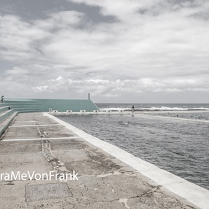 Könnte beinhalten: Ein Meerwasserpool mit helltürkisen Betontribünen und einem verwitterten Steindeck. Das Wasser ist dunkel und gekräuselt und trifft unter einem bewölkten Himmel auf den Horizont. Der Text FraMeVonFrank ist unten links in Weiß aufgedruckt.