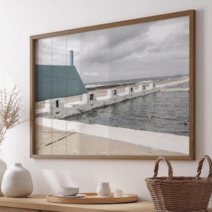 Könnte beinhalten: Gerahmter Druck eines Pools am Meer mit nummerierten Bahnen. Der Pool ist von weißem Beton umgeben und mit Wasser gefüllt. Im Hintergrund sind eine hellblaue Struktur und ein bewölkter Himmel zu sehen. Der Rahmen ist aus Holz.
