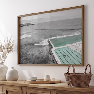 Könnte beinhalten: Ein gerahmter Druck einer Küstenlandschaft mit einem türkisfarbenen Schwimmbecken neben dem Meer. Das Bild ist überwiegend schwarz-weiß, wobei das Becken und ein kleiner Teil des Ozeans farbig sind. Der Rahmen ist hellbraun.