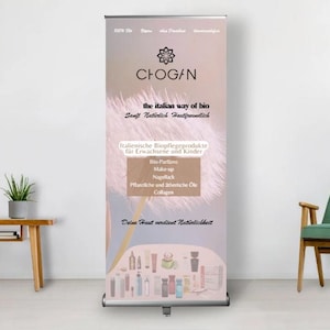 Könnte beinhalten: Ein vertikales Banner für Chogan, eine italienische Bio-Kosmetikmarke. Das Banner zeigt das Logo der Marke, Produktkategorien und den Slogan "the italian way of bio". Das Banner befindet sich in einem Raum mit einem kleinen Tisch, einem Stuhl und einer Topfpflanze.