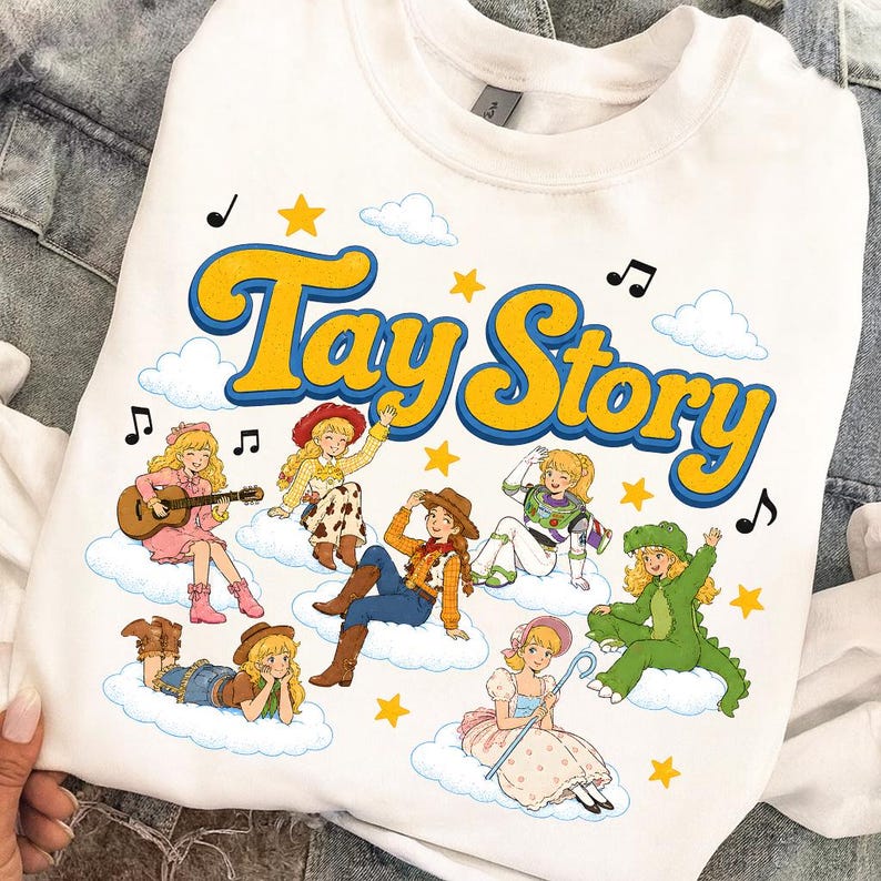 May include: A white crewneck sweatshirt featuring the text Tay Story in large yellow letters with blue outlines. The graphic includes multiple cartoon characters with blonde hair dressed in various costumes on clouds, surrounded by yellow stars and musical notes.