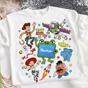 Op de afbeelding: Een wit t-shirt met een kleurrijke grafische print van Toy Story-personages, waaronder Woody, Buzz Lightyear, Jessie, Forky, Mr. Potato Head en een alien. Een centrale groene kikker houdt een blauw bord vast met de tekst Name.