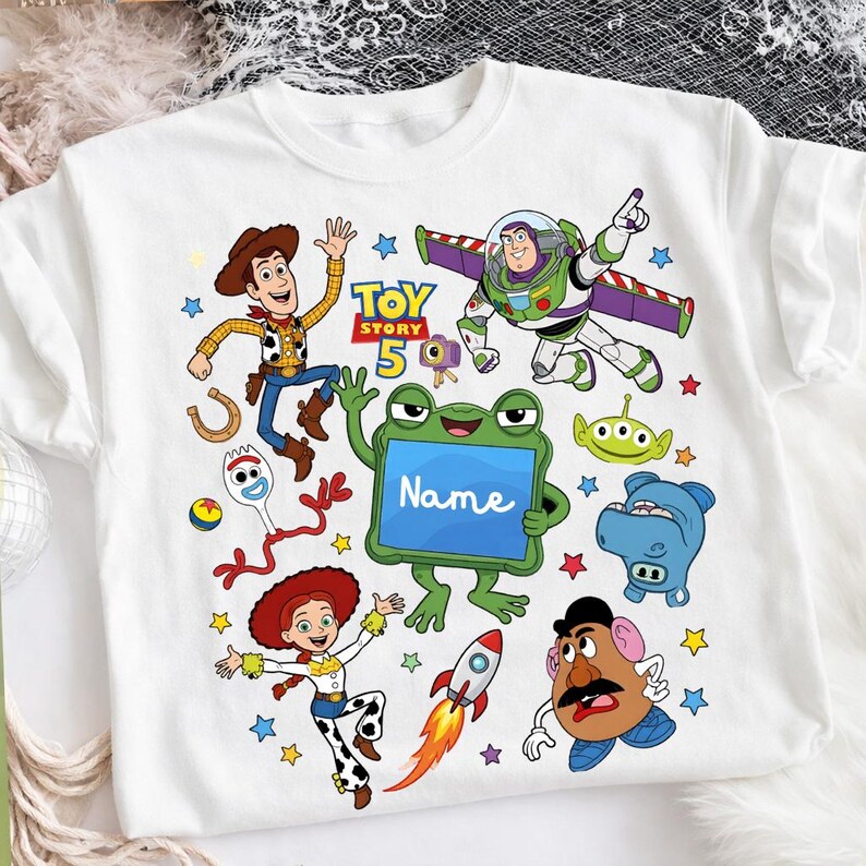 Op de afbeelding: Een wit t-shirt met een kleurrijke grafische print van Toy Story-personages, waaronder Woody, Buzz Lightyear, Jessie, Forky, Mr. Potato Head en een alien. Een centrale groene kikker houdt een blauw bord vast met de tekst Name.