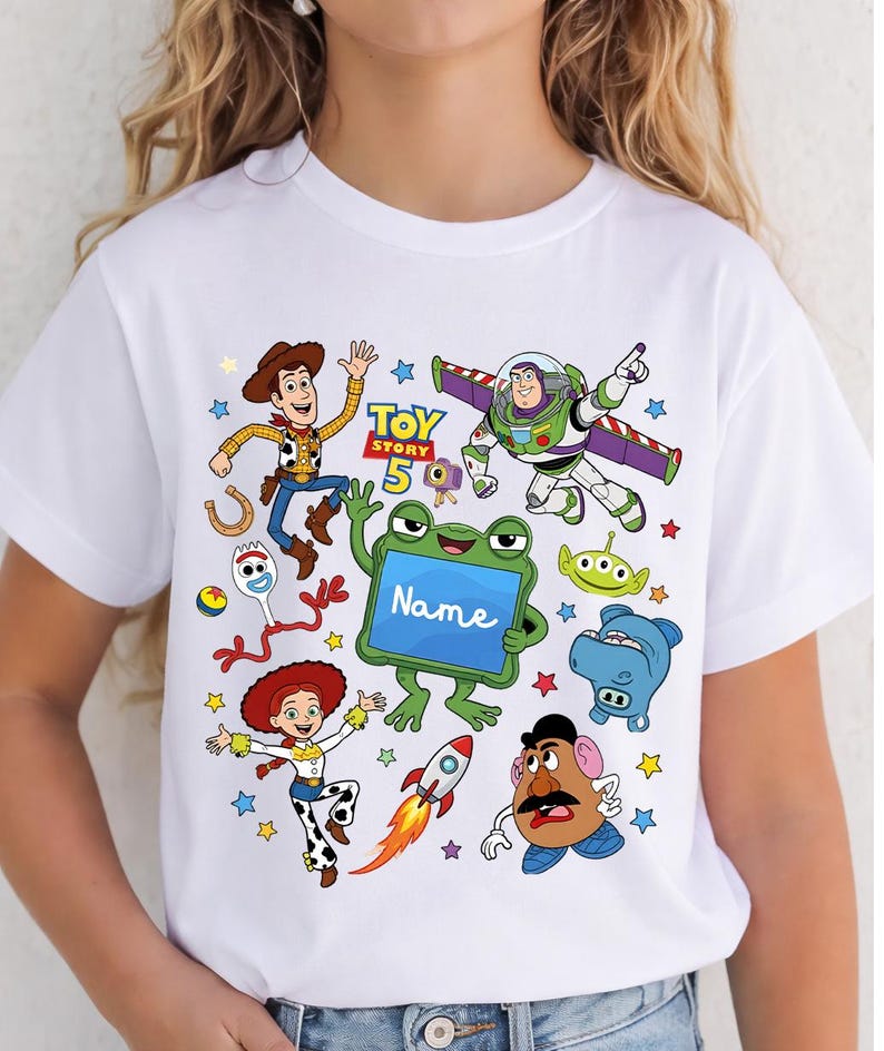 Op de afbeelding: Een wit t-shirt met een kleurrijke grafische print van Toy Story-personages, waaronder Woody, Buzz Lightyear, Jessie, Forky, Mr. Potato Head en een groene kikker die een blauw bord vasthoudt met de tekst Name. Kleine sterren en een raket zijn afgebeeld.