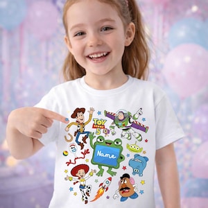 Op de afbeelding: Een wit t-shirt met een kleurrijke grafische print van Toy Story-personages zoals Woody, Buzz Lightyear, Jessie, Forky en Mr. Potato Head, met de tekst Toy Story 5 en een groene kikker die een bordje vasthoudt met de tekst Name.