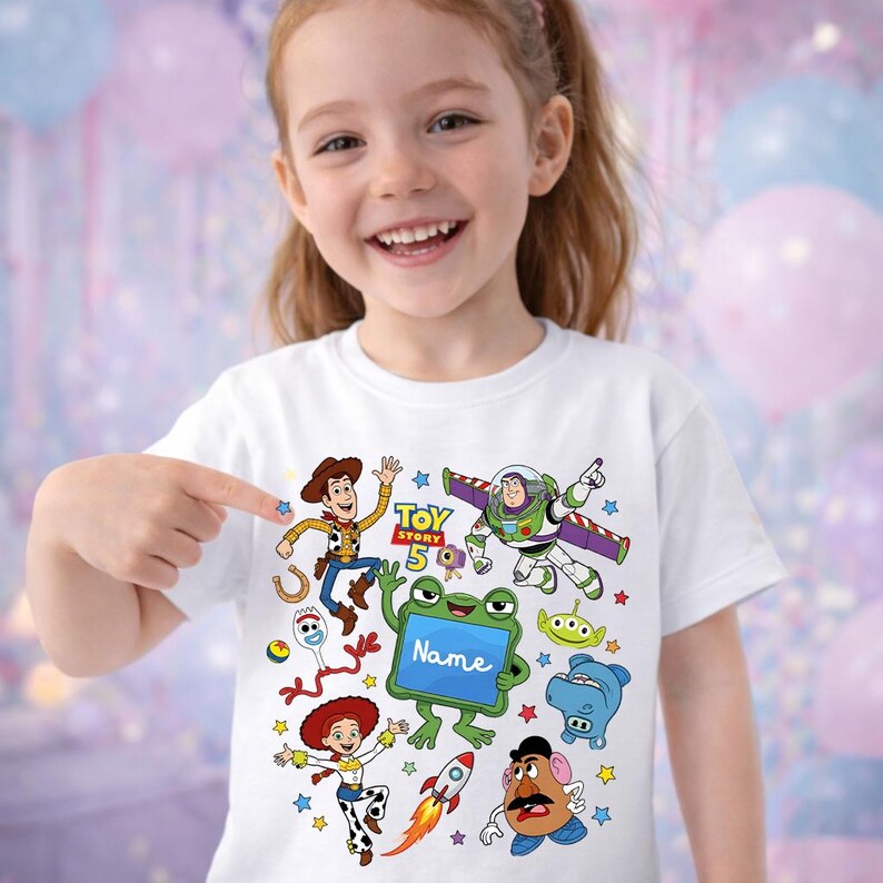 Op de afbeelding: Een wit t-shirt met een kleurrijke grafische print van Toy Story-personages zoals Woody, Buzz Lightyear, Jessie, Forky en Mr. Potato Head, met de tekst Toy Story 5 en een groene kikker die een bordje vasthoudt met de tekst Name.
