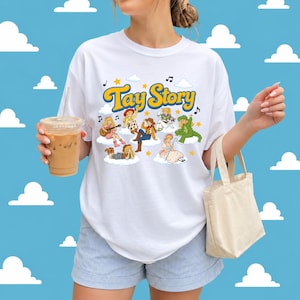 May include: A white short-sleeved t-shirt featuring a colourful graphic print with the text Tay Story in yellow letters. The design includes multiple cartoon characters dressed in various outfits, musical notes, and stars on a cloud background.