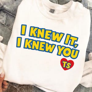 May include: A white crewneck sweatshirt featuring the yellow text with blue outlines that reads I KNEW IT, I KNEW YOU. Below the text is a red heart graphic containing the yellow letters TS with blue outlines, all printed on the front of the garment.