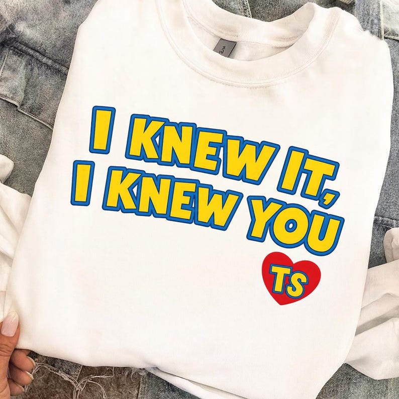 May include: A white crewneck sweatshirt featuring the yellow text with blue outlines that reads I KNEW IT, I KNEW YOU. Below the text is a red heart graphic containing the yellow letters TS with blue outlines, all printed on the front of the garment.