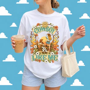 May include: A white t-shirt featuring a graphic print of two figures in cowboy hats, one playing a guitar, surrounded by orange flames, flowers, and a sun. The text "COWBOY LIKE ME" is printed in a teal, stylized font above and below the illustration.