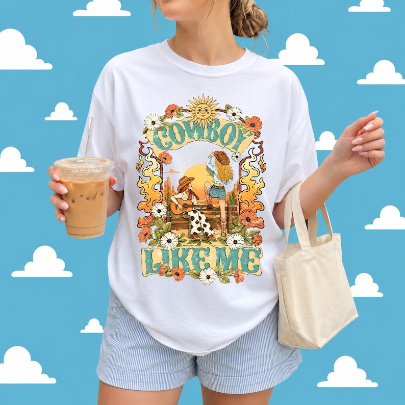 May include: A white t-shirt featuring a graphic print of two figures in cowboy hats, one playing a guitar, surrounded by orange flames, flowers, and a sun. The text "COWBOY LIKE ME" is printed in a teal, stylized font above and below the illustration.