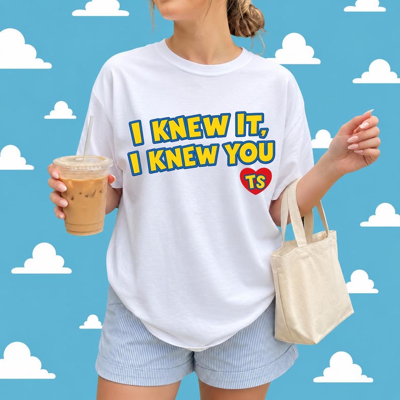 May include: A white short-sleeved t-shirt featuring the yellow text I KNEW IT, I KNEW YOU with a blue outline and a red heart containing the yellow letters TS. The shirt is paired with light blue and white pinstriped shorts and a beige tote bag.