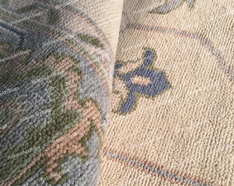May include: A close-up of a rolled-up rug with a beige background and a detailed pattern. The pattern includes blue, green, and pink floral and geometric designs. The rug's texture is visible, showing a dense weave. The brand name "Sabina Art Rugs" is visible.