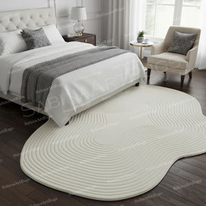 Op de afbeelding: Een crèmekleurig, abstract gevormd tapijt met een concentrisch, golfachtig patroon. Het tapijt ligt op een donkere houten vloer in een slaapkamer, gedeeltelijk onder een wit bed. De kamer omvat een stoel en een raam.