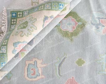 May include: A light blue rug with a floral pattern in shades of pink, green, and blue. The rug has a cream-colored border with a geometric design and fringe on one edge. The rug is folded over, revealing the underside.
