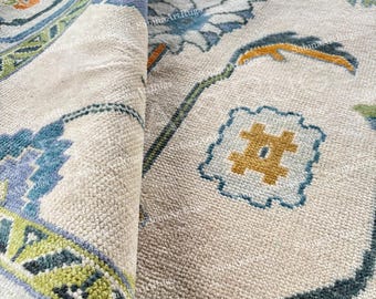 May include: Close-up of a beige rug with a detailed floral pattern in shades of blue, green, and orange. The rug is folded, revealing the intricate design and texture. The rug's design includes geometric shapes and floral motifs.