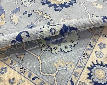 May include: A detailed close-up of a light blue rug with a rolled edge, showcasing intricate floral patterns in shades of blue and cream. The rug features a central medallion design and a border with repeating floral motifs. The rug is from SabinaArtRugs.