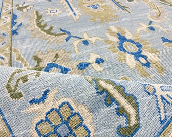 May include: A light blue rug with a detailed floral pattern in shades of blue, green, and cream. The rug has a rolled edge, revealing the textured weave and intricate design. The rug's design is a traditional style.