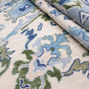 以下が含まれることがあります： 花柄の丸まったラグのクローズアップ。ラグは、クリーム色の背景に青と緑の花柄のデザインが施されています。ラグの質感が確認でき、丸まった部分はラグの厚みを示しています。