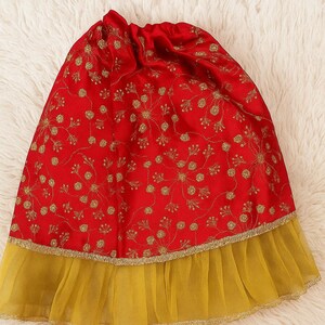 May include: A red skirt with gold floral embroidery and a yellow tulle hem. The skirt is made of a shiny fabric and has a drawstring waist. The skirt is likely for a child or a doll.