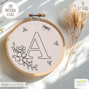 May include: An embroidery hoop featuring the letter "A" in black, surrounded by floral and dragonfly designs. The text "PDF PATTERN 6 SIZES" and "26 LETTERS" are visible. The design is on a cream-colored fabric, set in a wooden hoop.