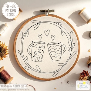 May include: Embroidery hoop with a cream-colored fabric featuring a black line drawing of two mugs, hearts, and a leafy wreath. The text "PDF + JPG PATTERN 6 SIZES" is in a white circle. The text "PRINTABLE A4 & LETTER SIZE" is at the bottom.