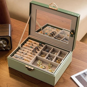May include: Open green jewelry box with a mirror inside, filled with rings, earrings, and other jewelry. The box is on a wooden surface. The box has a gold-colored clasp and handle.